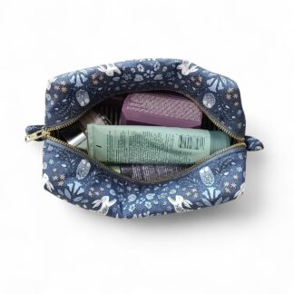 a-dark-blue-max-&-milo-toiletry-bag-or-cosmetic-pouch-with-a-nature-inspired-pattern-featuring-white-rabbits-flowers-and-foliage-shown-from-a-top-down-angle-with-a-visible-brass-YKK-zipper-displayed-with-cosmetic-products-inside